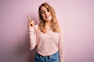 Young beautiful blonde woman wearing casual sweater and sunglasses over pink background showing and pointing up with finger number one while smiling confident and happy.