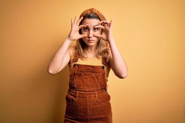 Young beautiful blonde woman wearing overalls and diadem standing over yellow background Trying to open eyes with fingers, sleepy and tired for morning fatigue