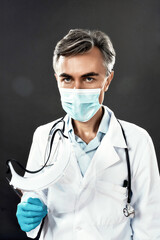 Stop Coronavirus. Mature male doctor in medical mask and blue gloves holding face shield, looking at camera while standing against dark background. Focus on man