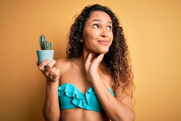 Young beautiful american woman on vacation wearing bikini holding cactus plant pot serious face thinking about question, very confused idea