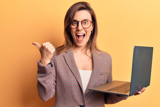 Young beautiful woman holding laptop pointing thumb up to the side smiling happy with open mouth