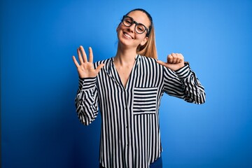 Beautiful blonde woman with blue eyes wearing striped shirt and glasses over blue background showing and pointing up with fingers number six while smiling confident and happy.