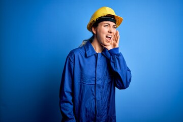 Young beautiful worker woman with blue eyes wearing security helmet and uniform shouting and screaming loud to side with hand on mouth. Communication concept.