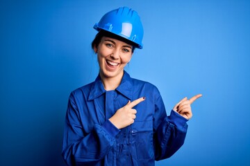 Young beautiful worker woman with blue eyes wearing security helmet and uniform smiling and looking at the camera pointing with two hands and fingers to the side.