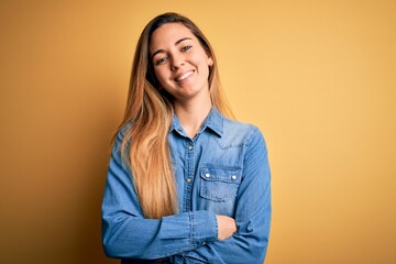 Fototapeta premium Young beautiful blonde woman with blue eyes wearing denim shirt over yellow background happy face smiling with crossed arms looking at the camera. Positive person.