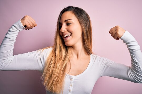 Young beautiful blonde woman with blue eyes wearing white t-shirt over pink background showing arms muscles smiling proud. Fitness concept.