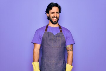 Young handsome hispanic clenaer man wearing housework apron and washing gloves winking looking at...