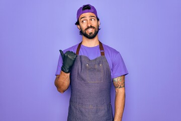 Young handsome hispanic man with bear wearing professional apron working as tattoo artist smiling with happy face looking and pointing to the side with thumb up.