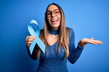 Young blonde woman with blue eyes holding colon cancer awareness blue ribbon very happy and...