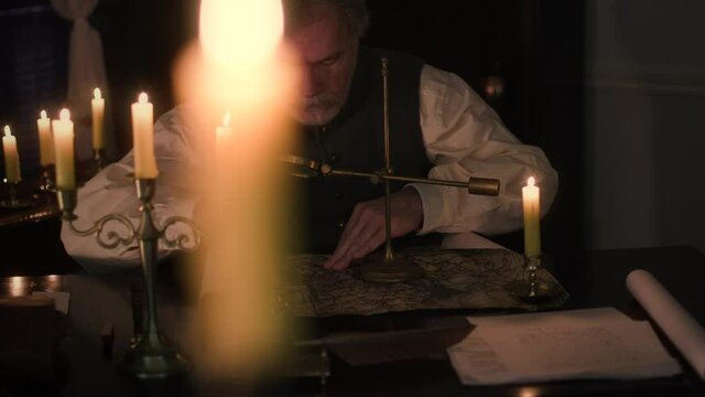 Tracking past an out of focus candle in the foreground to reveal a scene from the 18th century of an older man lit by candlelight using a magnifying glass to examine a map.