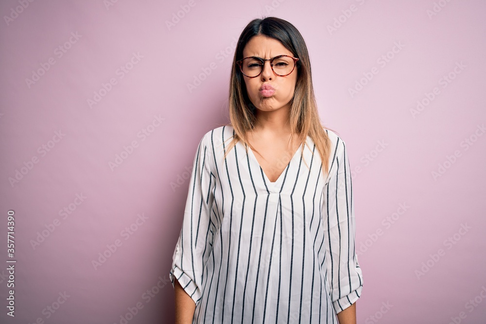 Sticker young beautiful woman wearing casual striped t-shirt and glasses over pink background puffing cheeks