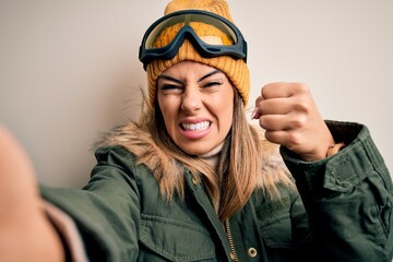 Young brunette skier woman wearing snow clothes and ski goggles make selfie by camera annoyed and frustrated shouting with anger, crazy and yelling with raised hand, anger concept