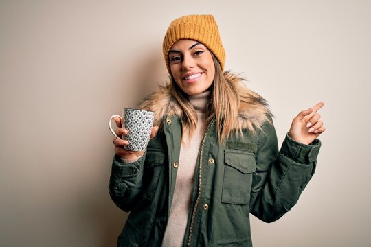 Young beautiful brunette woman wearing snow clothes drinking mug of coffee very happy pointing with hand and finger to the side