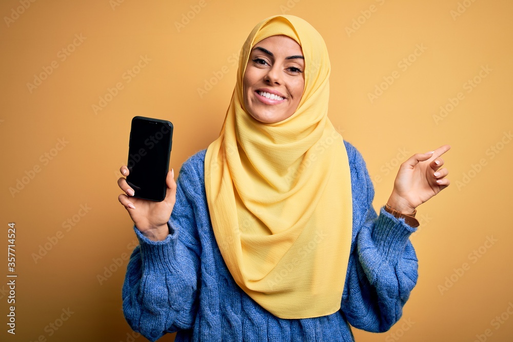 Poster Young beautiful brunette arab woman wearing islamic hijab holding smartphone showing screen very happy pointing with hand and finger to the side