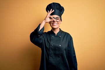 Young beautiful brunette chef woman wearing cooker uniform and hat over yellow background doing ok gesture with hand smiling, eye looking through fingers with happy face.