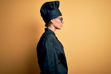 Young beautiful brunette chef woman wearing cooker uniform and hat over yellow background looking to side, relax profile pose with natural face with confident smile.