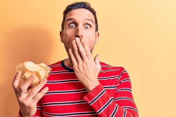 Young handsome man holding bowl of potato chips standing over isolated yellow background covering mouth with hand, shocked and afraid for mistake. Surprised expression