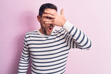 Young handsome man wearing casual striped sweater standing over isolated pink background peeking in shock covering face and eyes with hand, looking through fingers afraid