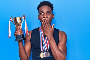 Young african american man holding champion trophy wearing medals covering mouth with hand, shocked...