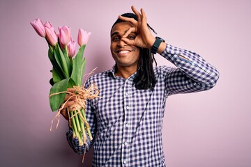 Young african american afro romantic man with dreadlocks holding bouquet of pink tulips with happy face smiling doing ok sign with hand on eye looking through fingers