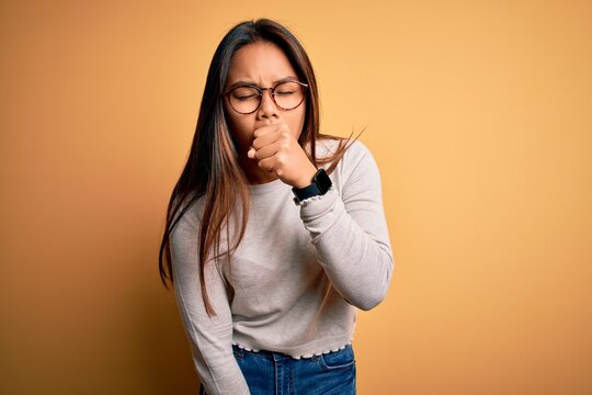 Young beautiful asian girl wearing casual sweater and glasses over yellow background feeling unwell and coughing as symptom for cold or bronchitis. Health care concept.