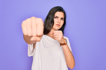 Young beautiful brunette woman wearing casual white t-shirt over purple background Punching fist to fight, aggressive and angry attack, threat and violence