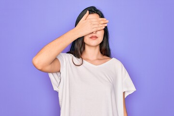 Young beautiful brunette woman wearing casual white t-shirt over purple background covering eyes with hand, looking serious and sad. Sightless, hiding and rejection concept