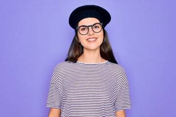 Young beautiful brunette woman wearing glasses and french beret over purple background with a happy and cool smile on face. Lucky person.