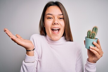 Young beautiful brunette girl holding small cactus plant pot over isolated white background very happy and excited, winner expression celebrating victory screaming with big smile and raised hands