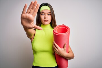 Beautiful sporty girl doing sport wearing sportswear holding yoga mat over white background with open hand doing stop sign with serious and confident expression, defense gesture