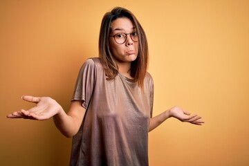 Beautiful brunette girl wearing casual t-shirt and glasses over isolated yellow background clueless and confused expression with arms and hands raised. Doubt concept.