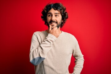 Young handsome man with beard wearing casual sweater standing over red background with hand on chin thinking about question, pensive expression. Smiling with thoughtful face. Doubt concept.