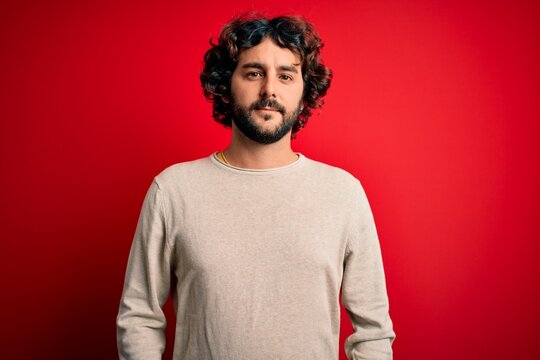 Young handsome man with beard wearing casual sweater standing over red background Relaxed with serious expression on face. Simple and natural looking at the camera.