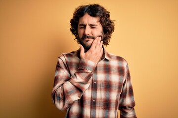 Fototapeta premium Young handsome man with beard wearing casual shirt standing over yellow background touching mouth with hand with painful expression because of toothache or dental illness on teeth. Dentist