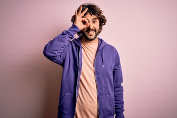 Young handsome sporty man with beard wearing casual sweatshirt over pink background doing ok gesture with hand smiling, eye looking through fingers with happy face.