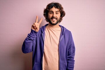 Young handsome sporty man with beard wearing casual sweatshirt over pink background showing and pointing up with fingers number two while smiling confident and happy.