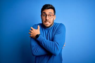 Young handsome man with beard wearing casual sweater and glasses over blue background shaking and freezing for winter cold with sad and shock expression on face