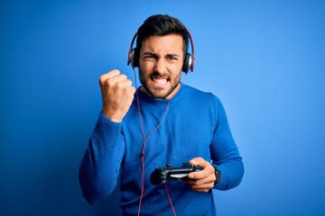 Young handsome gamer man with beard playing video game using joystick and headphones annoyed and...