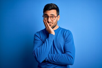 Young handsome man with beard wearing casual sweater and glasses over blue background thinking...