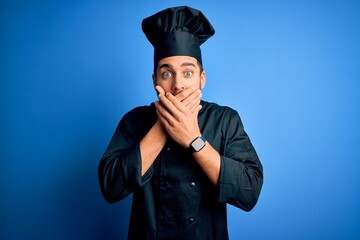 Young handsome chef man with beard wearing cooker uniform and hat over blue background shocked covering mouth with hands for mistake. Secret concept.