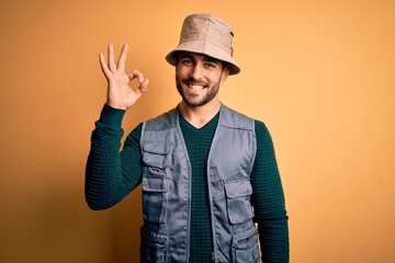 Handsome tourist man with beard on vacation wearing explorer hat over yellow background smiling positive doing ok sign with hand and fingers. Successful expression.