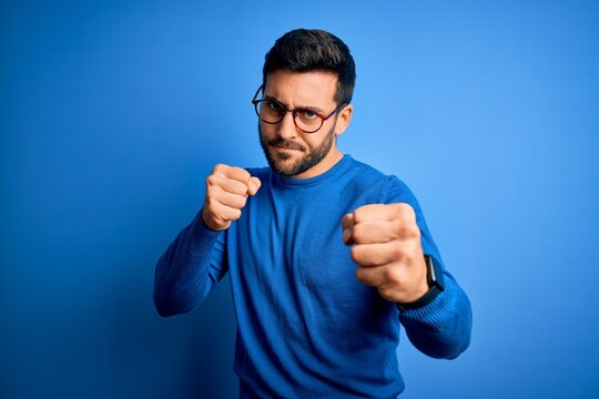 Young handsome man with beard wearing casual sweater and glasses over blue background Punching fist to fight, aggressive and angry attack, threat and violence