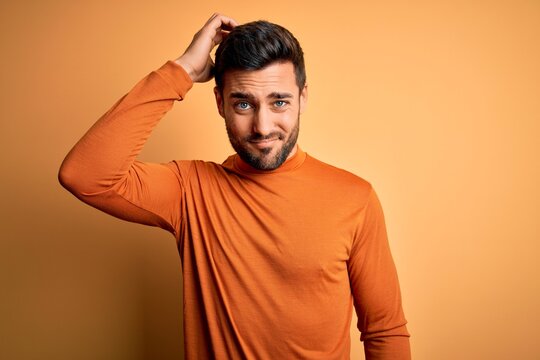 Young handsome man with beard wearing casual sweater standing over yellow background confuse and wonder about question. Uncertain with doubt, thinking with hand on head. Pensive concept.