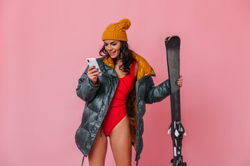 Curious girl in swimsuit using smartphone. Studio shot of pretty young woman in down jacket with skis.