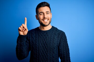 Young handsome man wearing casual sweater standing over isolated blue background showing and pointing up with finger number one while smiling confident and happy.