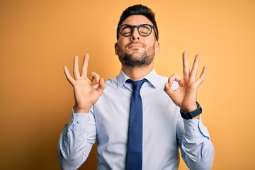 Young handsome businessman wearing tie and glasses standing over yellow background relaxed and smiling with eyes closed doing meditation gesture with fingers. Yoga concept.