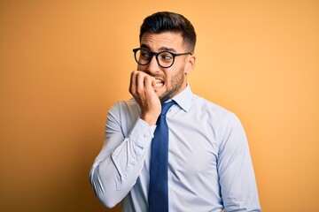 Young handsome businessman wearing tie and glasses standing over yellow background looking stressed and nervous with hands on mouth biting nails. Anxiety problem.