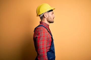 Young builder man wearing construction uniform and safety helmet over yellow isolated background looking to side, relax profile pose with natural face with confident smile.