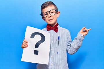 Cute blond kid wearing nerd bow tie and glasses holding question mark smiling happy pointing with hand and finger to the side