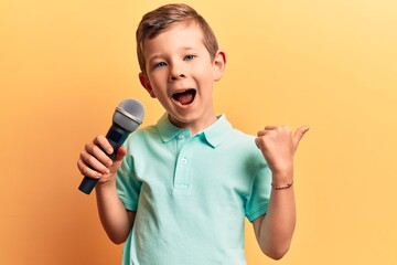 Cute blond kid singing song using microphone pointing thumb up to the side smiling happy with open...